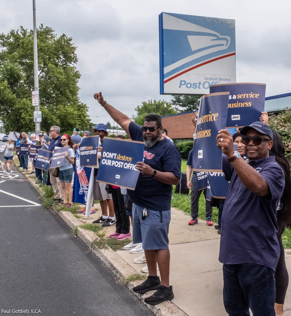 A group of people holding signs outside a post office with a big USPS sign above them.